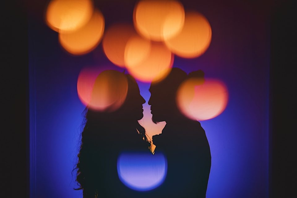 Silhouettes of a couple facing each other against a vibrant blue background with soft, orange bokeh light effects at the Ash + John Boathouse wedding.