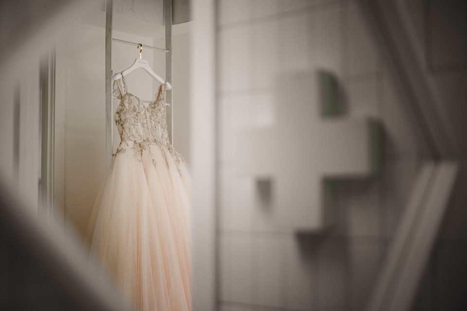An elegant peach-colored bridal gown with intricate lace details hangs on a hanger in a softly lit boathouse, with a reflective surface and blurred background.