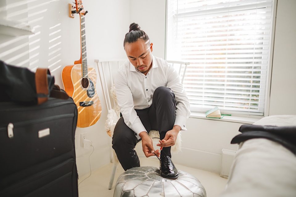 A man ties the laces of his black shoes in a bright boathouse, with a guitar hanging on the wall and luggage nearby.