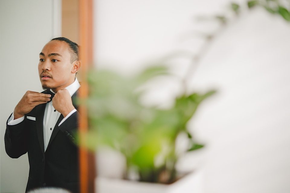 A man in a tuxedo adjusting his bow tie while looking in the mirror, with a blurred indoor plant in the foreground at an Ash + John Boathouse NYC wedding.