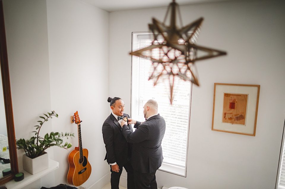 A person helps adjust the tie of another person in a tuxedo at Ash + John Boathouse NYC Wedding in a well-lit room featuring a guitar, window, and hanging star lamp.
