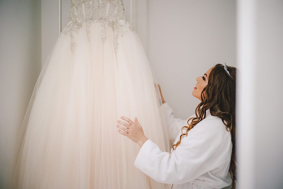 Bride in a white robe admiring her hanging wedding dress at the NYC Wedding, touching it gently with both hands.