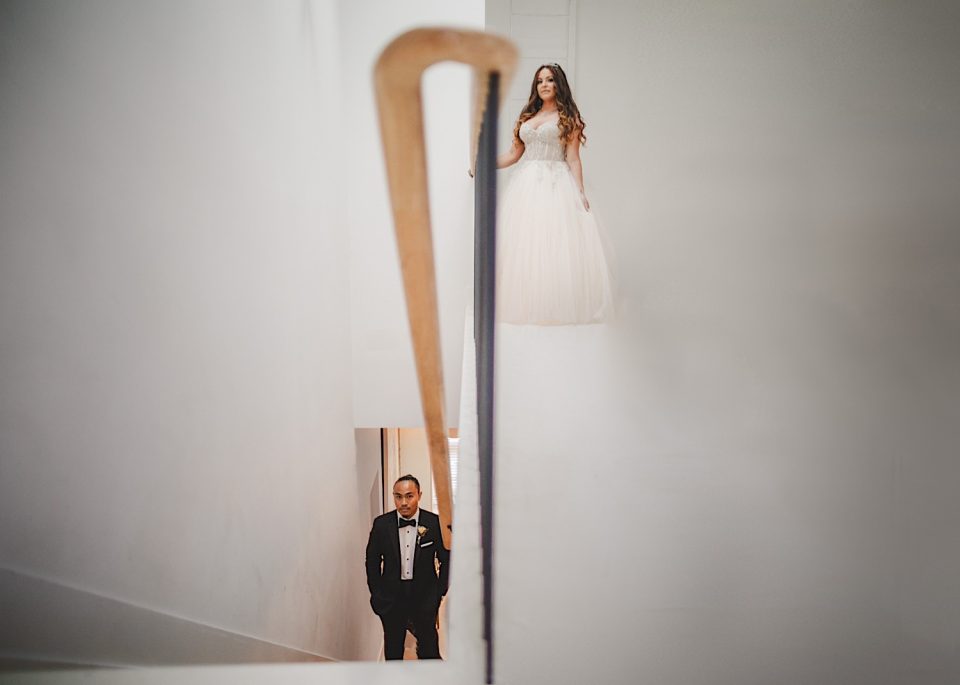 A bride and groom seen through a doorway at the Ash + John Boathouse; the bride stands at the top of stairs while the groom looks up from below.
