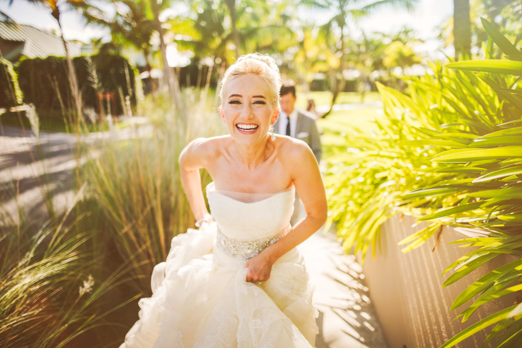 A bride in a strapless white dress smiles widely while walking outdoors on a sunny day with greenery around her. A groom follows behind, capturing the perfect moment for their wedding photography.