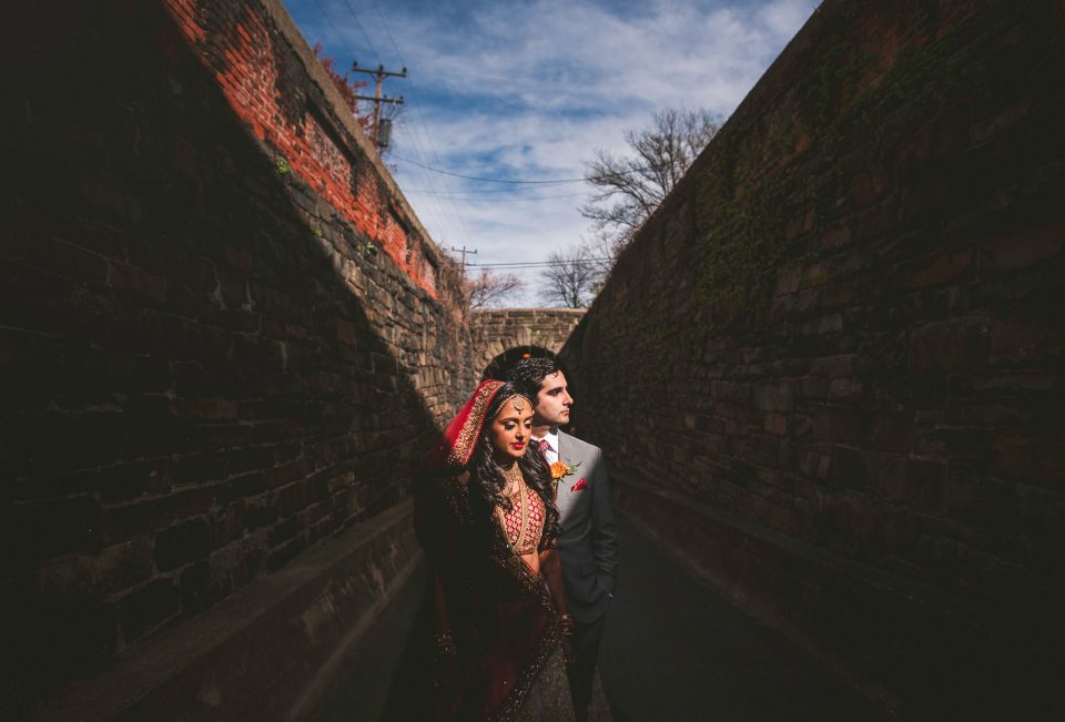 A bride in traditional attire and a groom in a suit stand together in a narrow, shaded alleyway with brick walls under a blue sky, perfectly capturing the essence of timeless wedding photography.