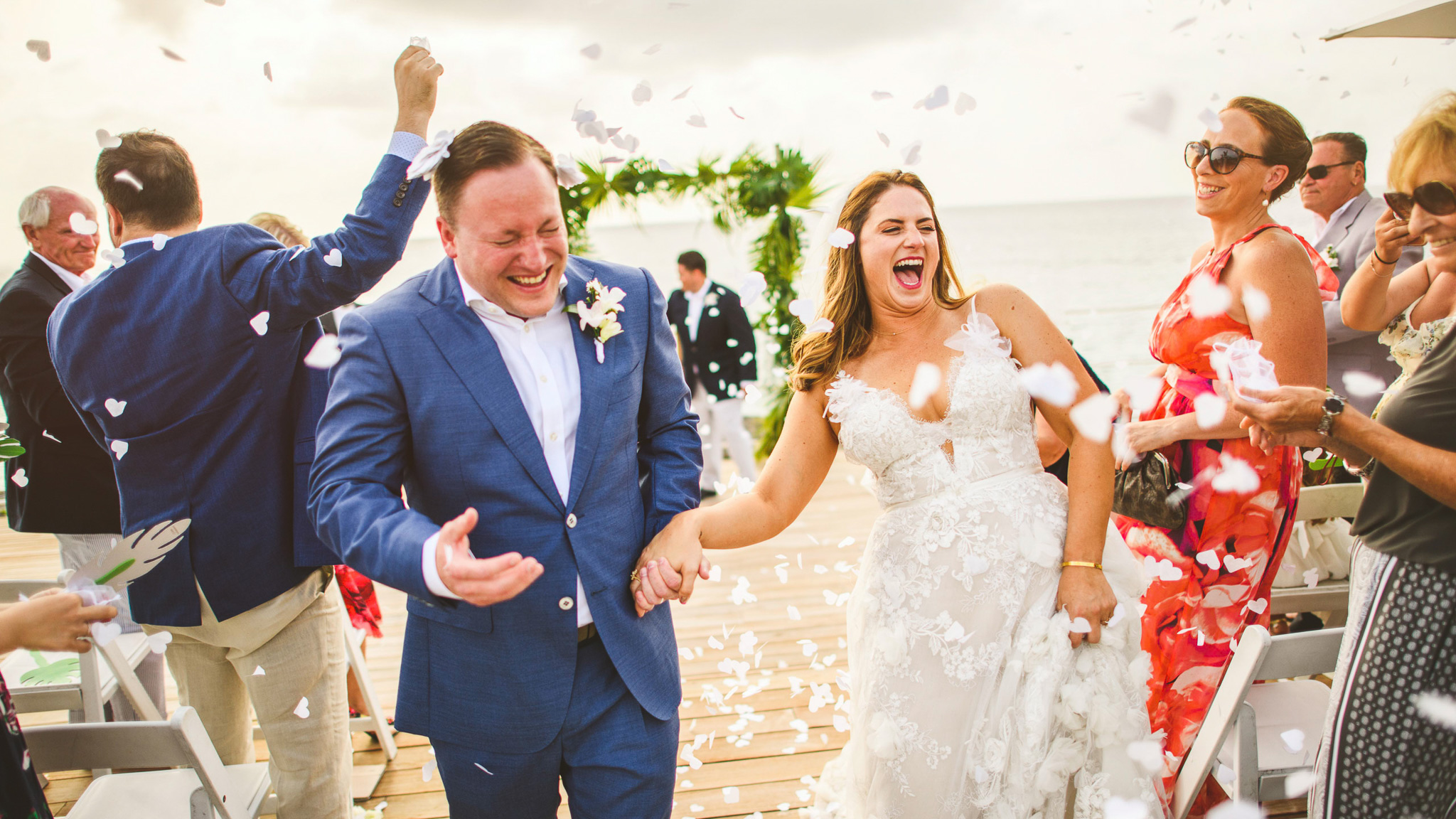 A bride and groom walk down the aisle, smiling and laughing as guests throw white confetti, beautifully captured by expert wedding photography. The woman wears a lace wedding gown, and the man is in a blue suit. The background shows an outdoor setting. For premium pricing details, contact us.