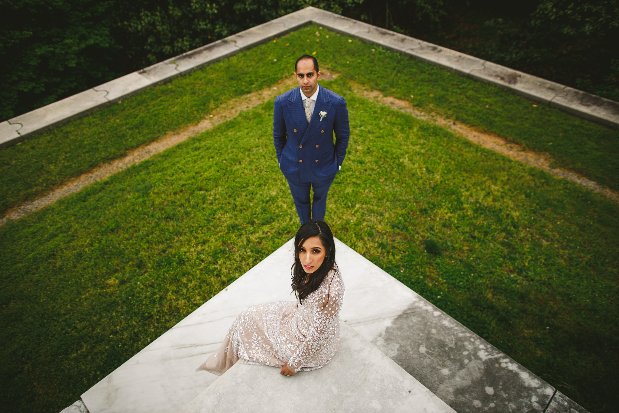 A woman in a light-colored dress sits on a concrete surface, while a man in a blue suit stands on grass behind her. Positioned in a triangular arrangement with greenery in the background, this photograph captures the essence of wedding elegance.