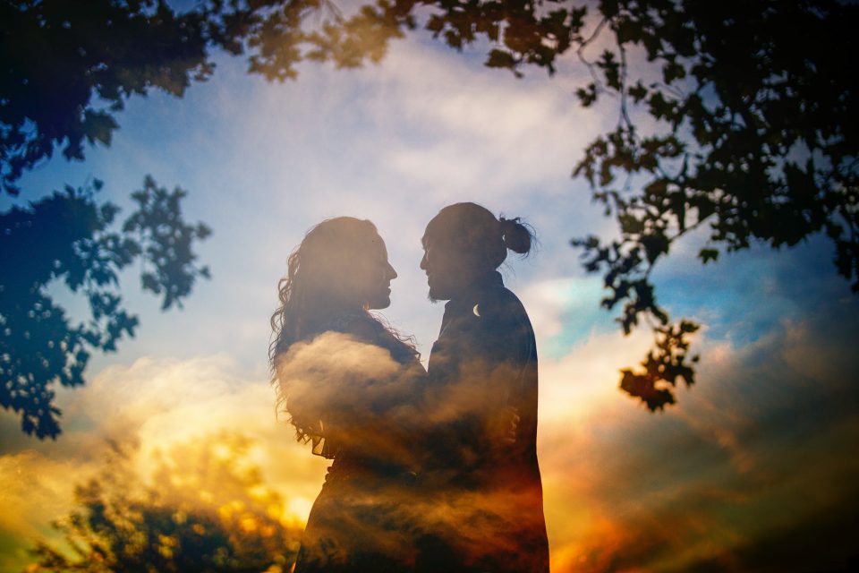 Silhouette of two people standing face to face surrounded by a colorful, cloudy sky and tree branches in the foreground—a perfect moment for wedding photography.