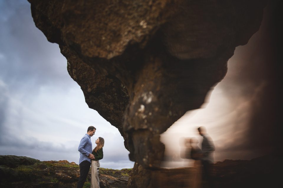 A couple stands near a majestic rock formation, their blurred reflection shimmering to the side under a moody, cloud-laden sky.