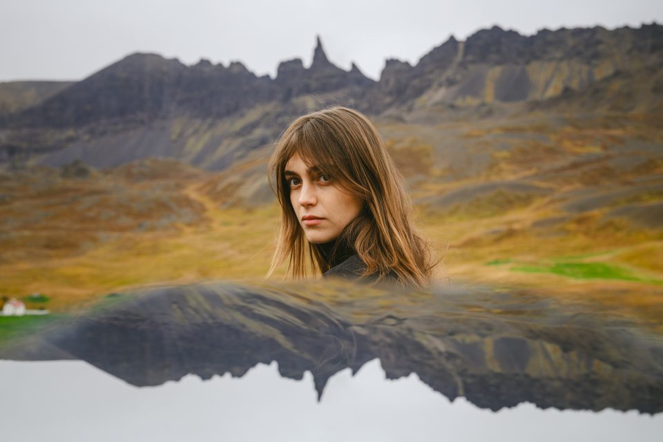 A person with long hair stands outdoors, framed by a blurred auto draft-like reflection in the foreground, with a breathtaking mountainous landscape in the background.