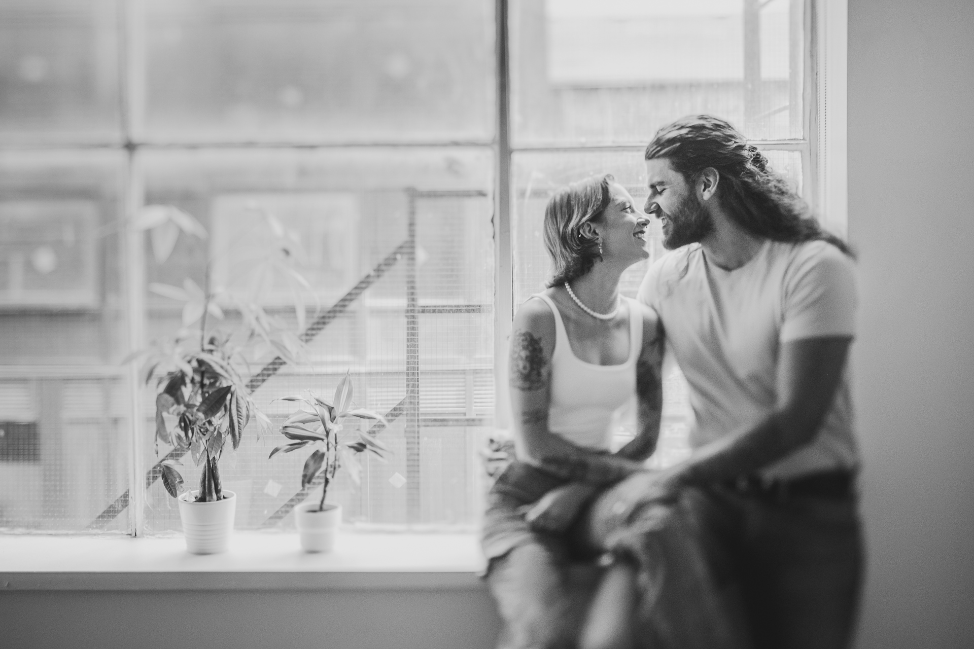 A black and white photo of a couple sitting on a window ledge, looking at each other and smiling. Two potted plants, adding a touch of nature to the scene, are positioned on the ledge beside them.