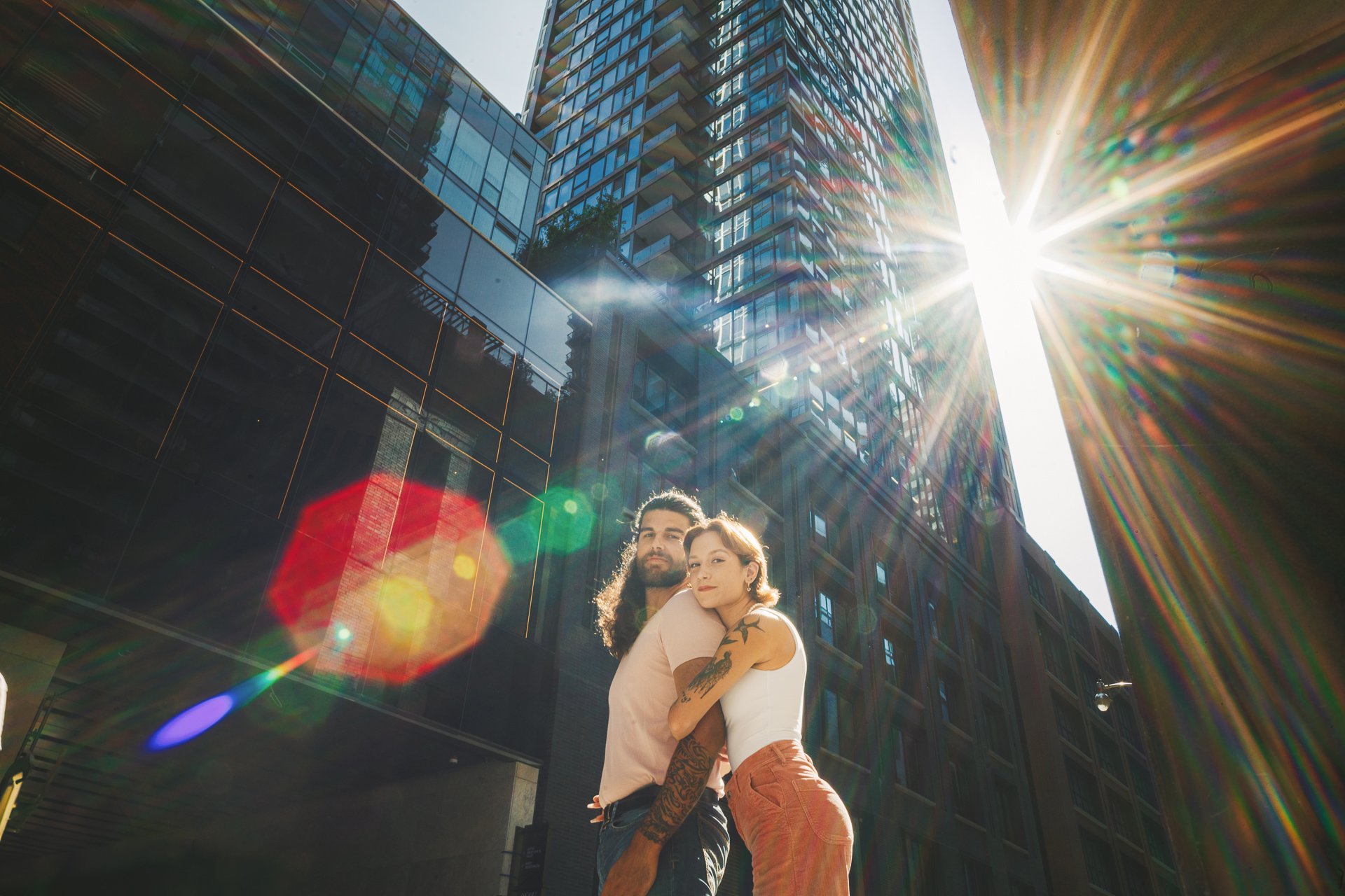 A bearded man and a tattooed woman stand close together on a city street with tall buildings and a bright sun flare in the background, creating an almost cinematic scene that could have been an Auto Draft from a modern love story.