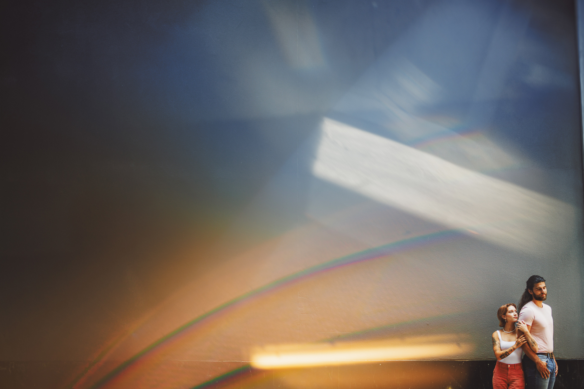 A man and woman stand close together against a large, dark wall with vibrant, rainbow-like light reflections, creating an almost auto draft of colorful hues.