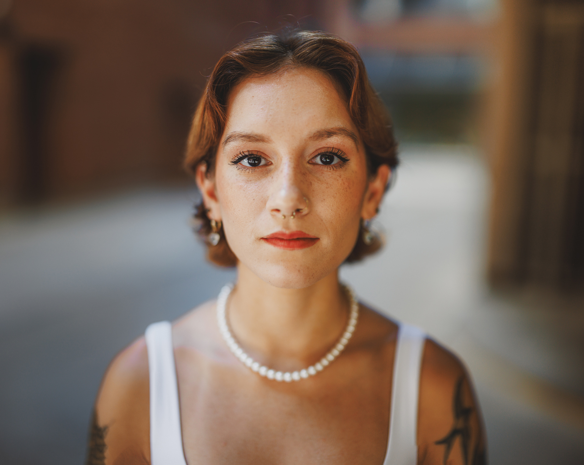 A woman with short brown hair and a white tank top looks directly at the camera, standing outdoors. She is wearing a pearl necklace and has visible tattoos on her arms, as if captured in an auto draft snapshot of a candid moment.