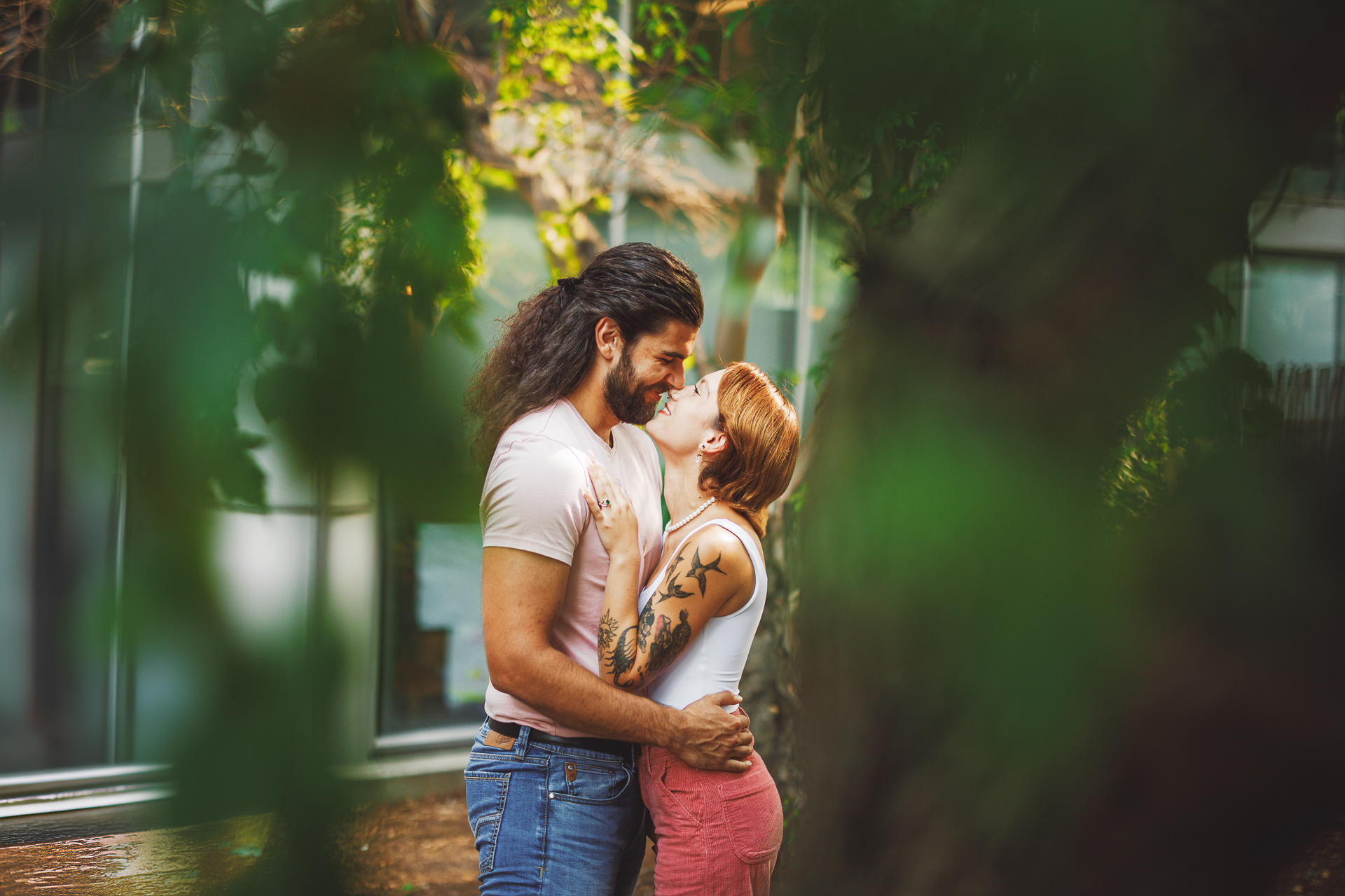 A man and woman stand close together outdoors, smiling at each other like an auto draft of love. The woman has her hands on the man's chest, and they are surrounded by greenery.