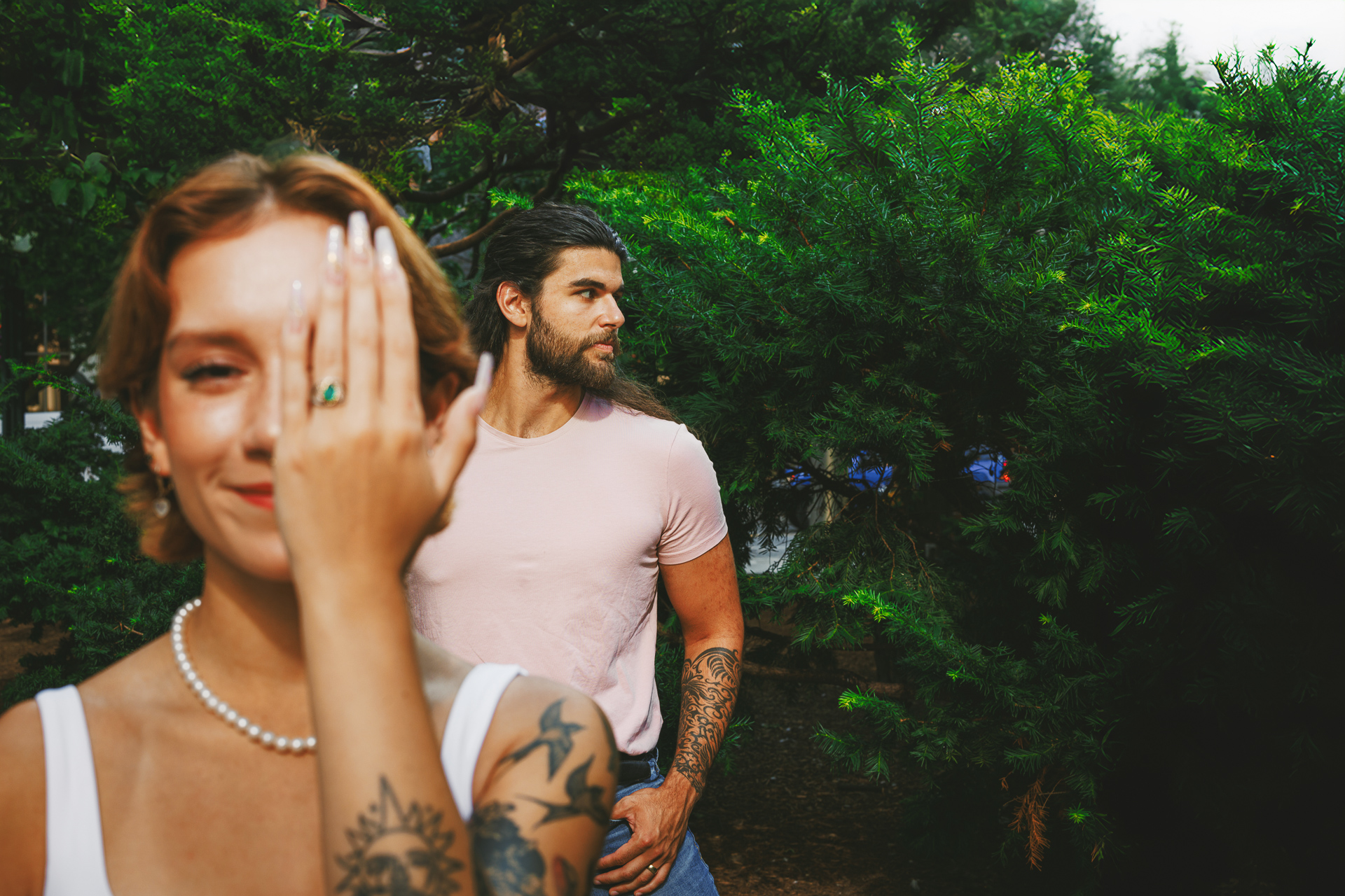 A tattooed woman shows off a draft engagement ring, covering one eye with her hand, while a man stands in the background against a lush backdrop of green bushes.