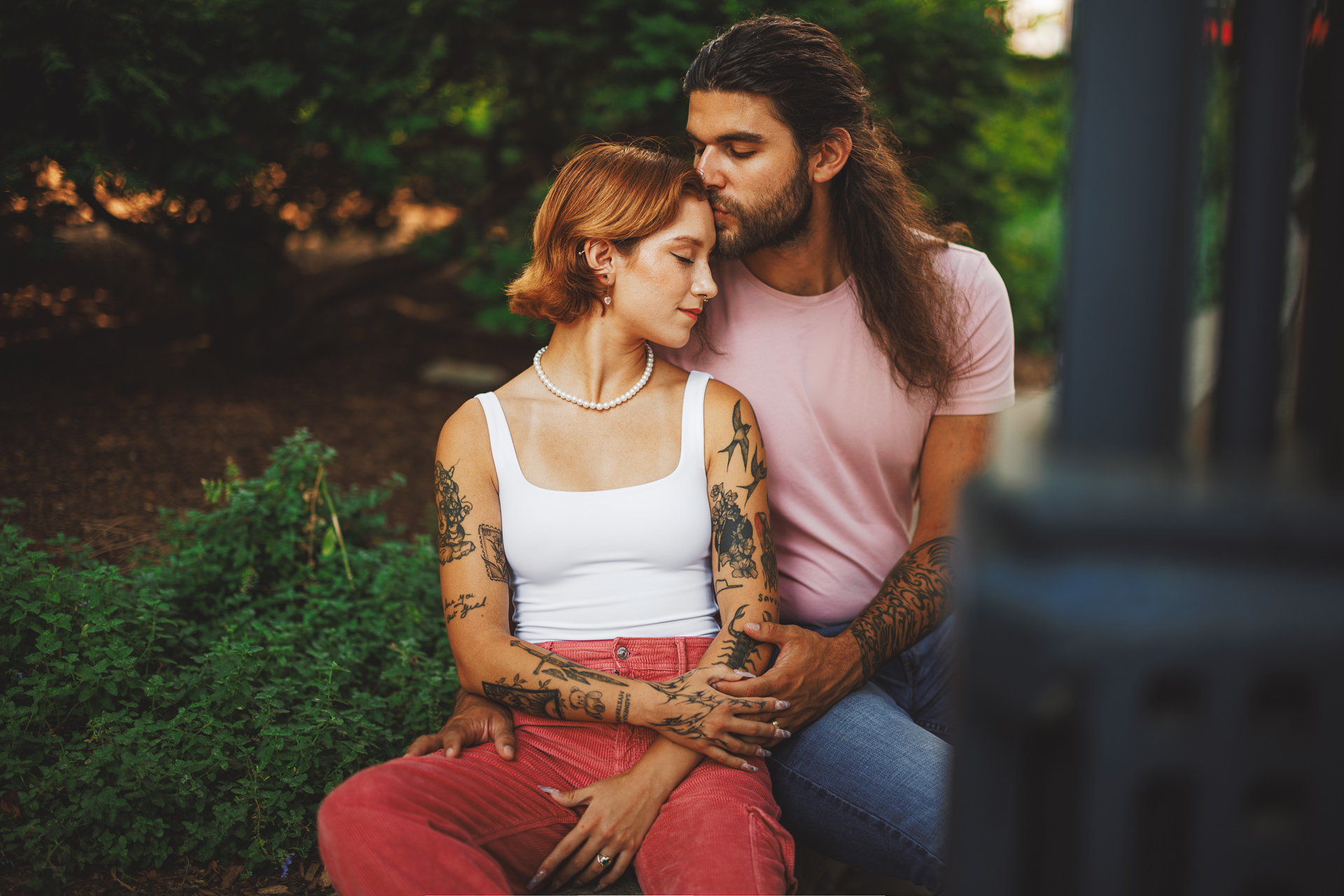 A man with long hair kisses the forehead of a woman with short hair as they sit close together in an outdoor setting, surrounded by greenery. She has multiple tattoos and wears a white tank top and pink pants, their moment seeming almost like it was captured by auto draft.