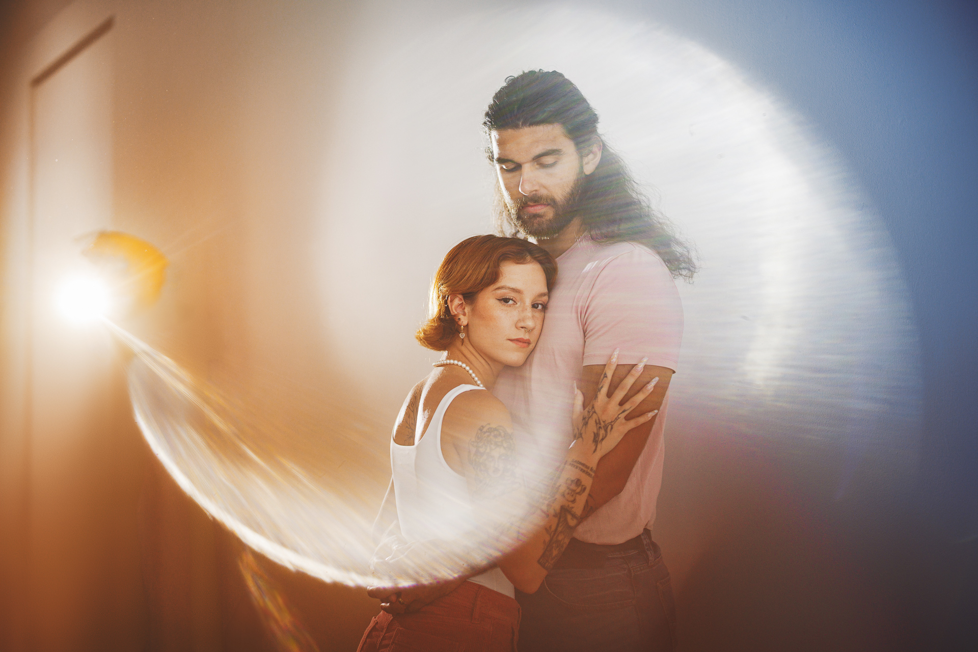 A man and woman stand closely together, with the woman leaning on the man. Light flares create a circular pattern around them, as if captured in an auto draft of their intimate moment.