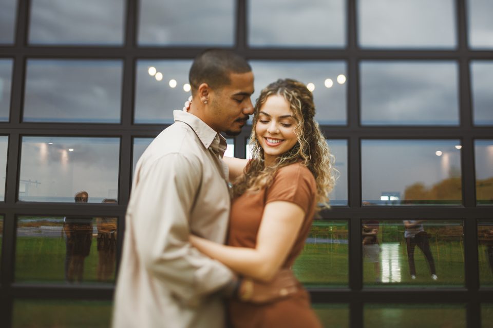 A couple embraces closely, smiling, in front of a large window reflecting the outdoors. The sky is overcast, and other people are faintly seen in the reflection, as if part of an auto draft sketch of life unfolding.