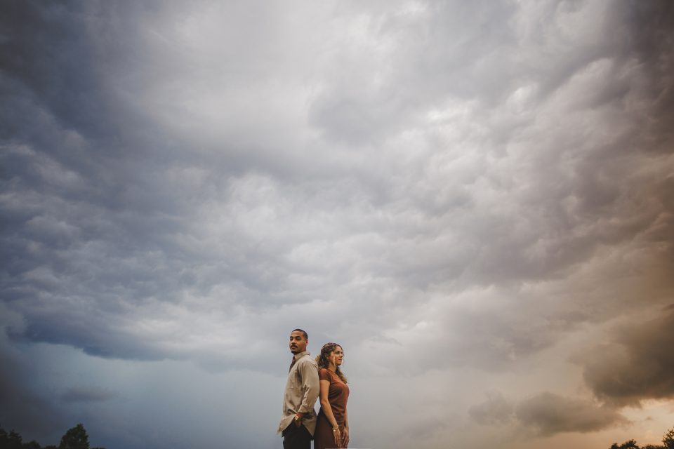 A man and a woman stand back-to-back under a dramatic, cloudy sky, looking in opposite directions, as if drafted into separate worlds. The landscape is largely covered by the vast clouds above.
