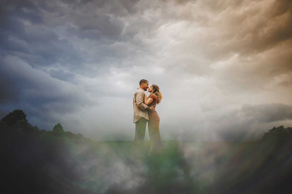 A couple stands closely on a foggy landscape under a dramatic cloudy sky, with mist and sunlight reflecting around them like an auto draft of ethereal beauty.