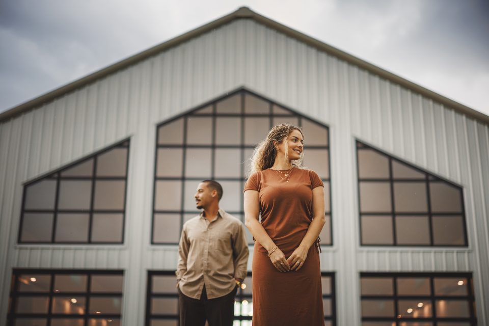 Two individuals, one in a brown dress and the other in a beige shirt, stand in front of a modern, large building with multiple glass windows. The woman is looking to the right, and the man to the left, as if poised for an auto draft of their next move.