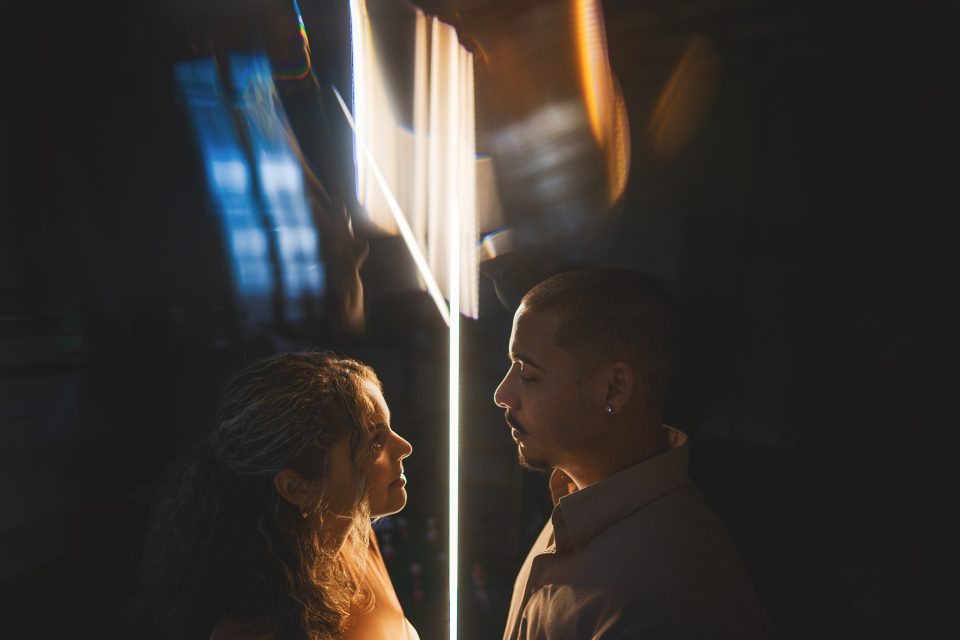 Two people facing each other closely in low light, with a vertical streak of bright light between them, casting dramatic shadows. The background is dark with blurred and abstract light reflections, almost like an auto draft of a moment in time.