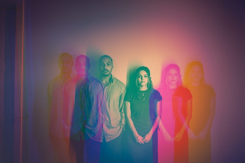 A man and a woman stand side by side against a wall, with a multicolored repetition effect giving the illusion of multiple overlapping images, like an auto draft of vibrant echoes.