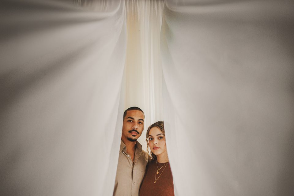 A man and a woman stand closely together, framed by white curtains, like an intimate moment captured in an auto draft.
