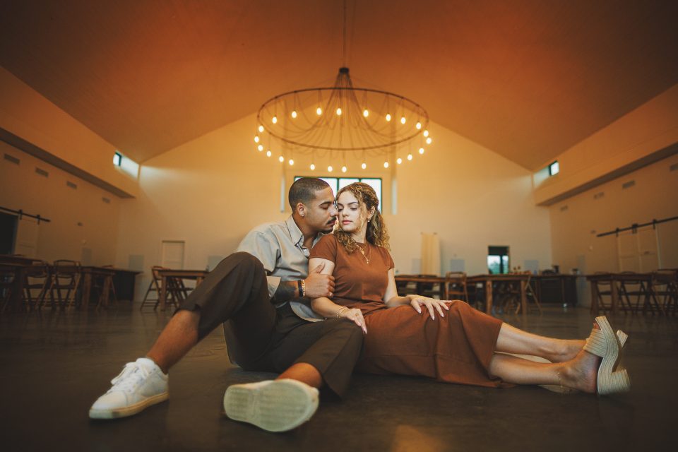A couple sits together on the floor of a large, empty room with a high, vaulted ceiling and a modern chandelier, as if waiting for their own story to auto draft into this serene space.