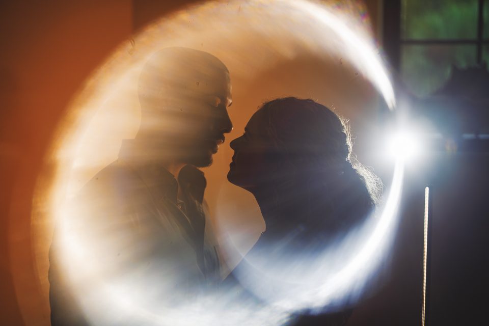 Silhouetted couple facing each other closely, surrounded by a halo of light in a dimly lit room, as if their moment was an auto draft sketch of pure intimacy.