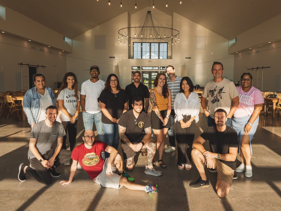 A group of thirteen people pose for a photo inside a spacious, well-lit hall adorned with chandeliers. Some are standing while others kneel or sit gracefully on the floor, creating a moment that seems almost like an auto draft of timeless elegance.