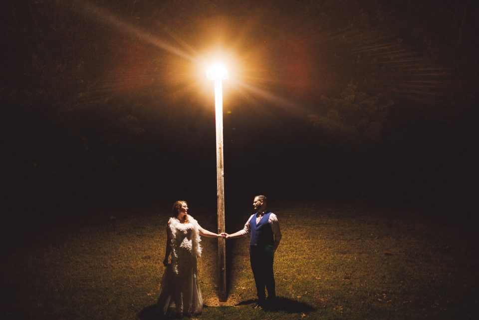 A bride and groom hold hands under a street lamp at night, standing on an otherwise dark lawn, as if captured in an auto draft of a timeless memory.