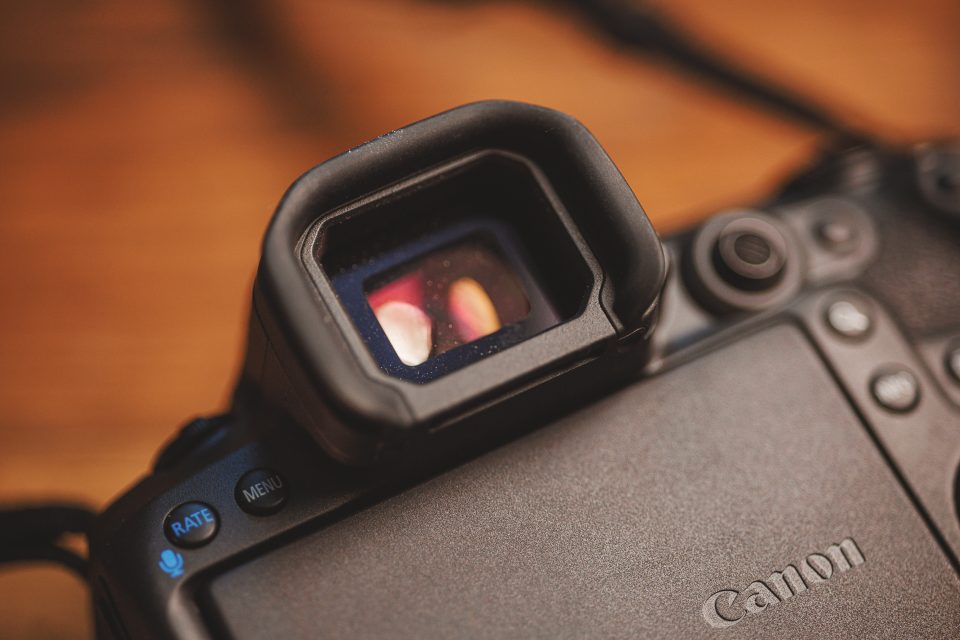 Close-up of a Canon R5 Mark II's viewfinder and rear buttons, including a blue microphone button and a rate button on the left. The background is blurred, capturing the essence of real-world use.