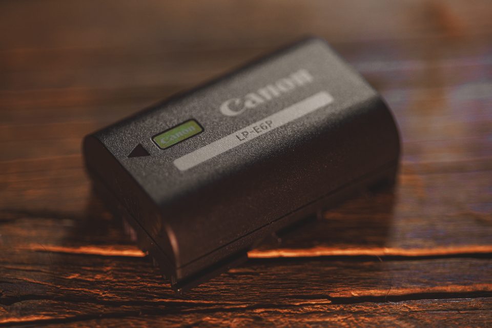 Close-up of a Canon LP-E6P camera battery on a wooden surface. The battery, used in the Canon R5 Mark ii for real-world use, is black with a green label and the Canon logo.