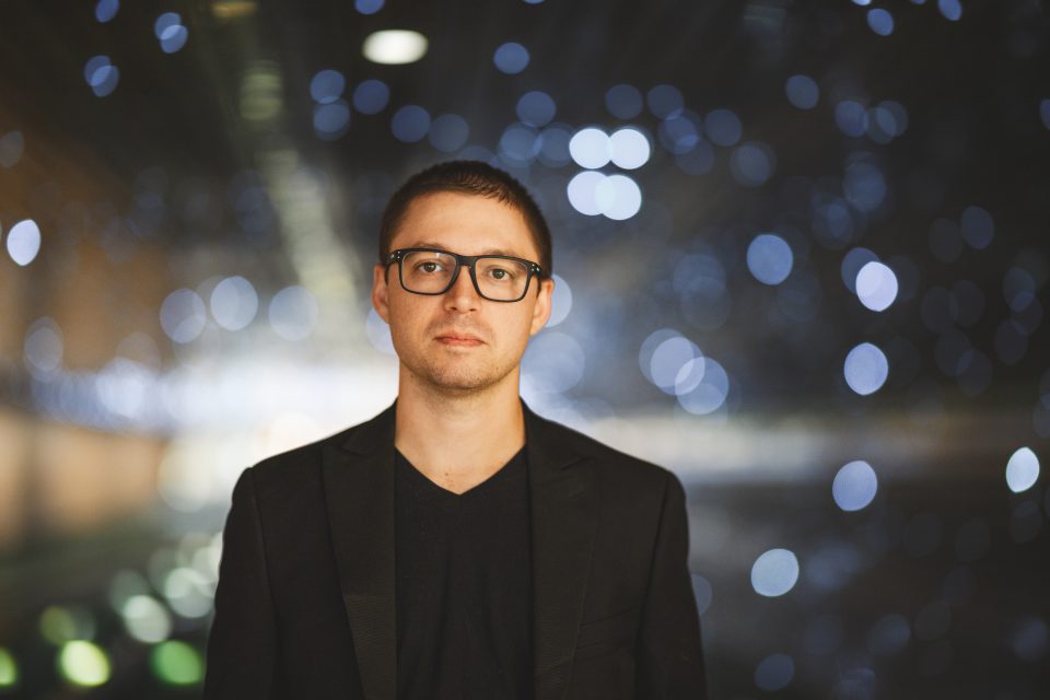 A person wearing glasses and a black blazer stands in front of a blurred background with various circular lights.