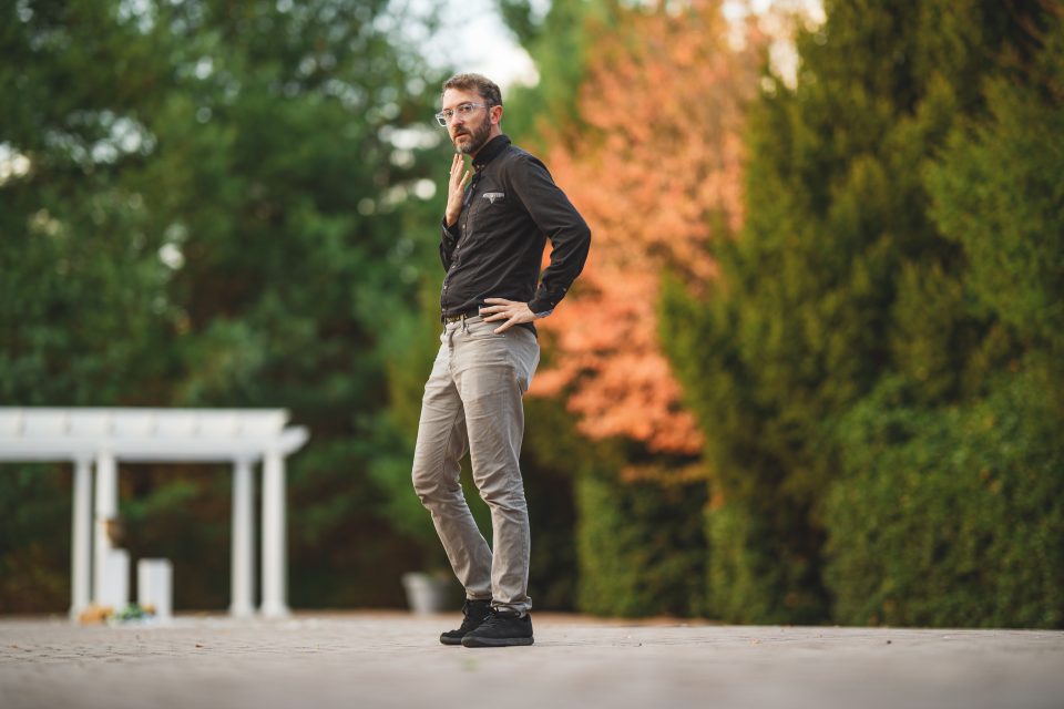 A person in casual attire stands outdoors with hands on hips, surrounded by greenery and autumn-colored trees.