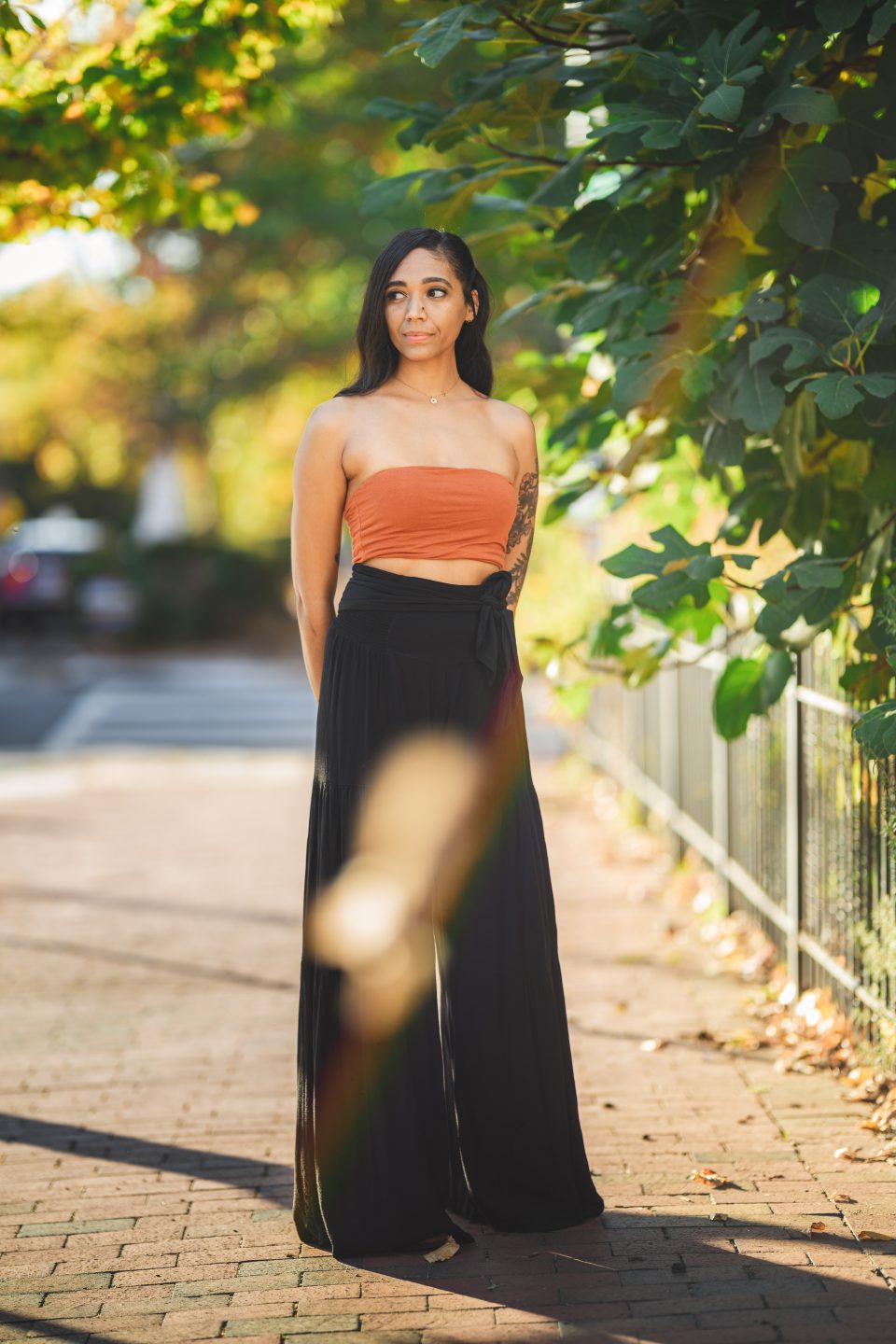 A woman stands on a sunlit brick path, wearing a strapless orange top and black pants, with greenery in the background.