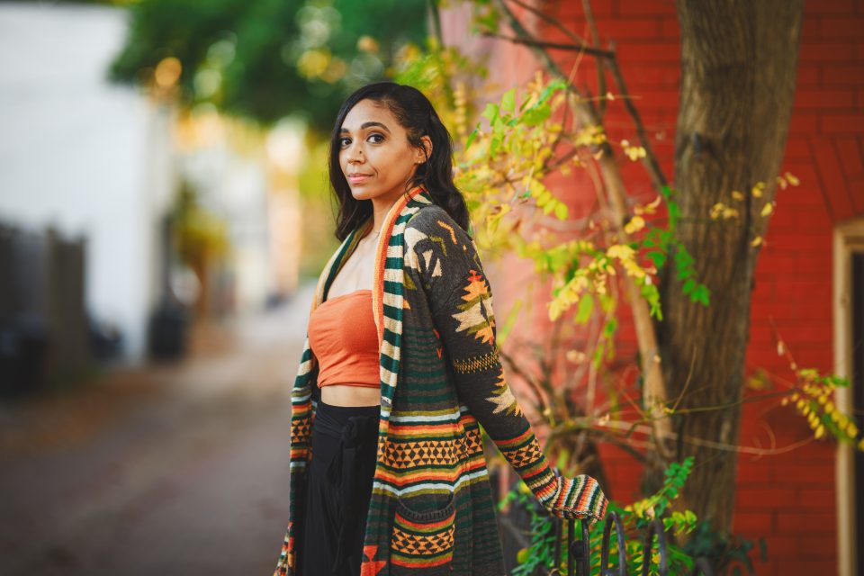 A woman in an orange top and patterned cardigan stands outdoors near a brick wall and tree, with colorful leaves in the background.