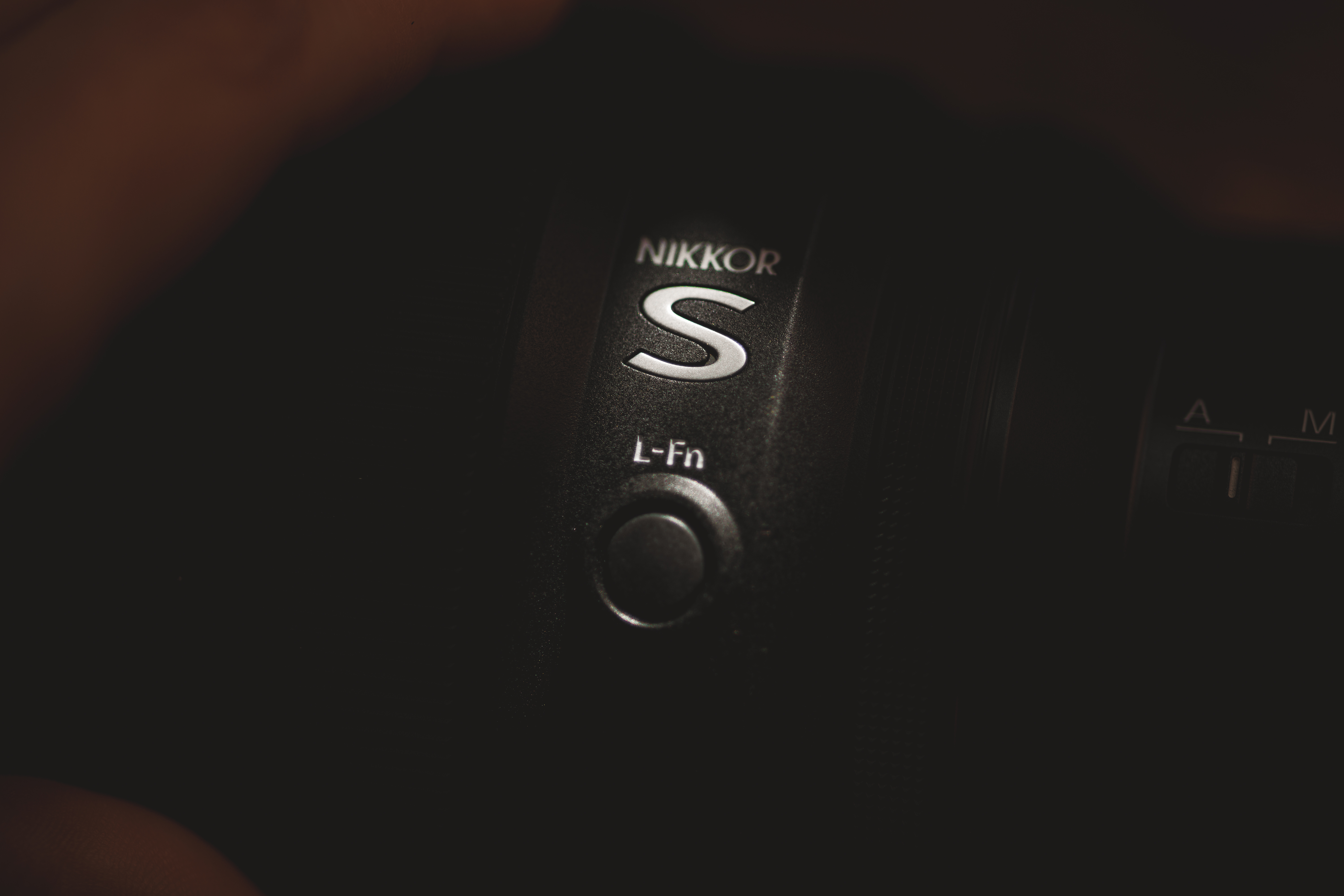 Close-up of a NIKKOR camera lens with the letter "S" and "L-Fn" button visible, partially illuminated in low light.