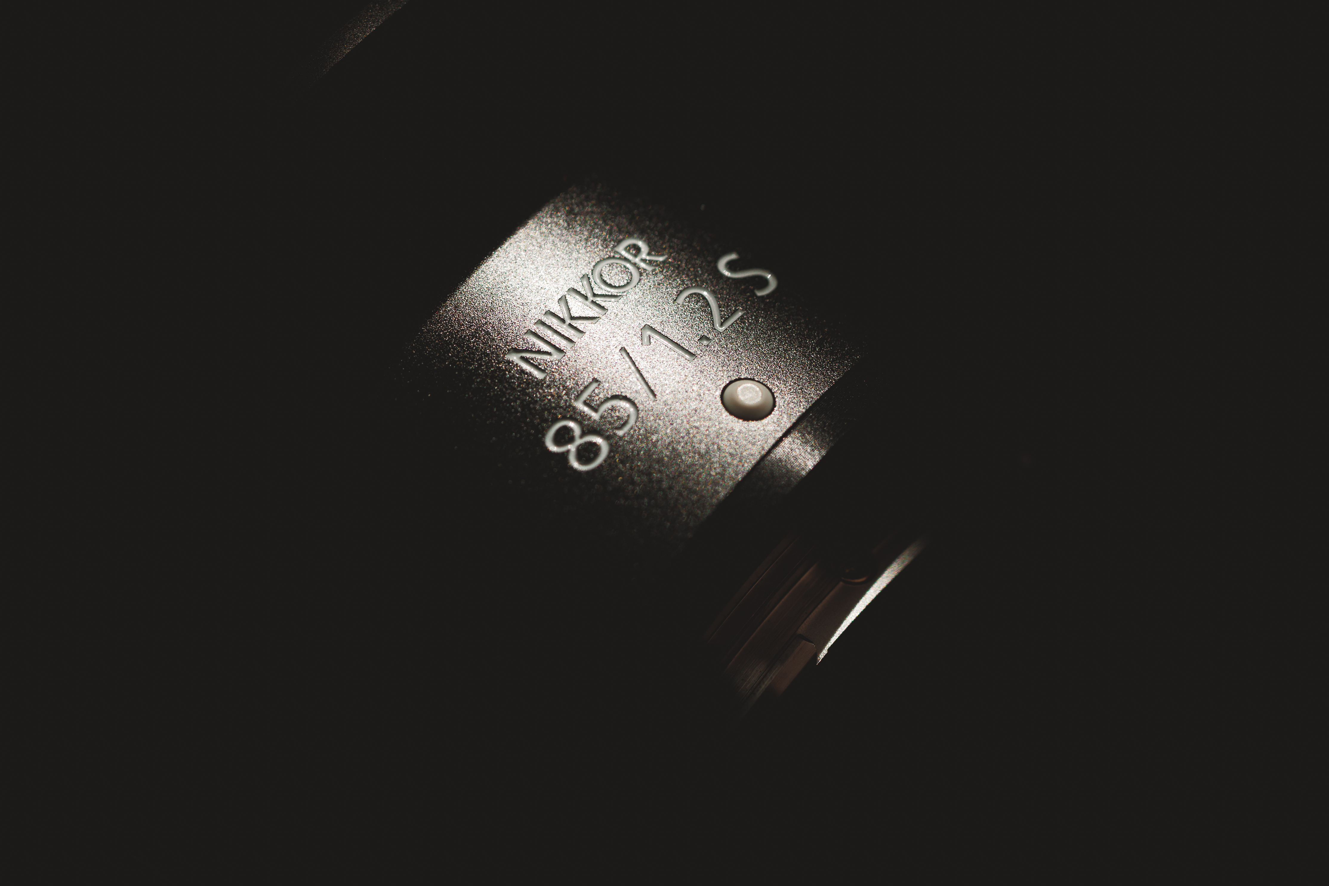 Close-up of a Nikkor 85mm f/1.2 S lens in low light, highlighting the engraved text on the lens barrel.