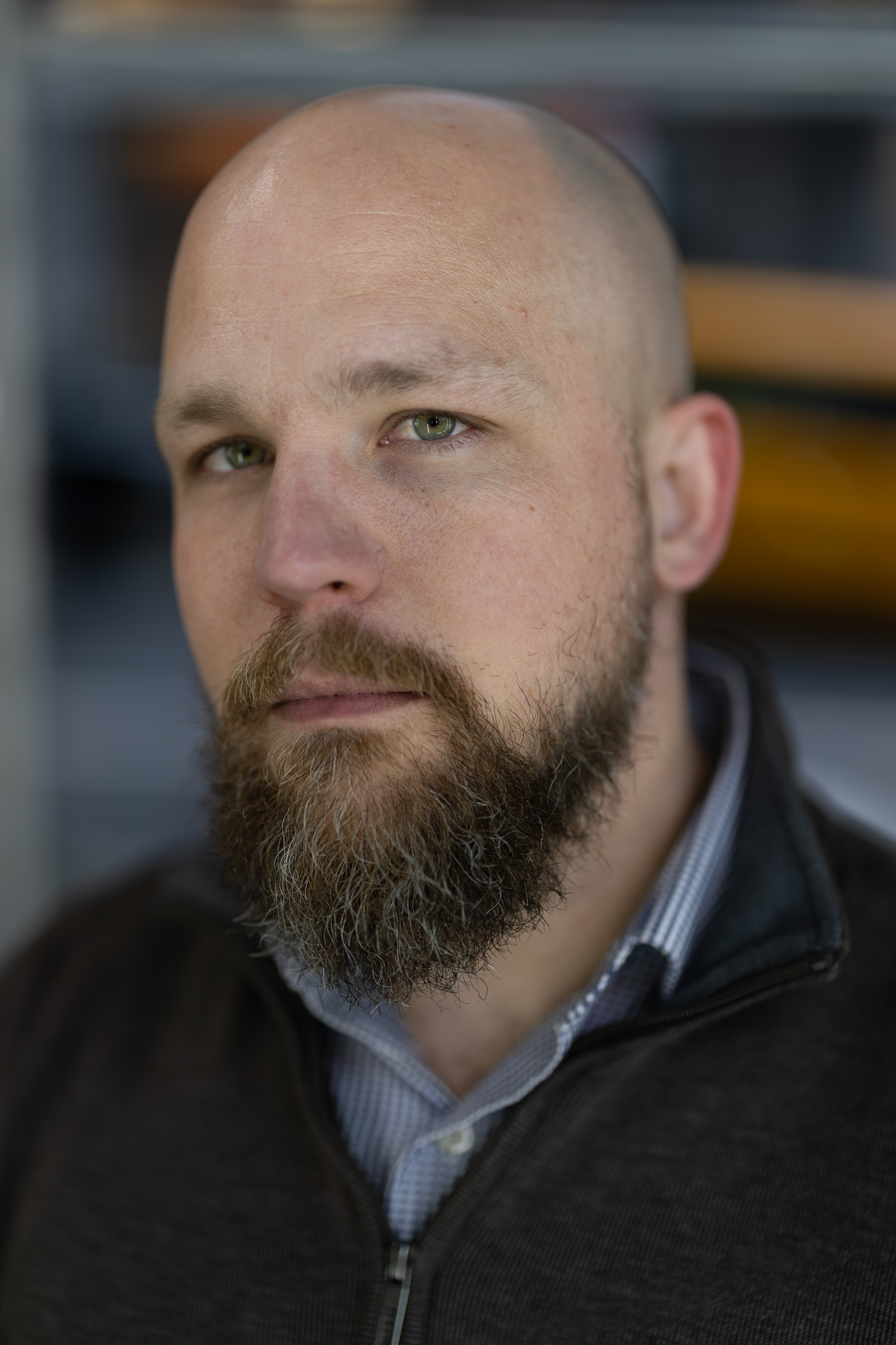 A bearded man with a serious expression looks into the camera, reminiscent of someone about to review their work. He is bald and wears a dark sweater over a collared shirt, as if ready for an intense session of AI editing with AfterShoot.
