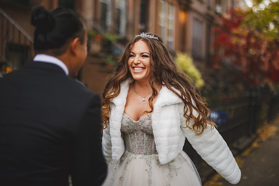 A bride in a white fur jacket and tiara laughs joyfully while facing a man in a suit, with autumn leaves falling during their NYC wedding.