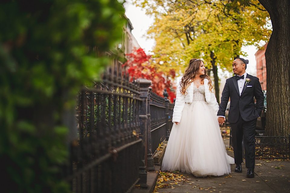 A bride and groom walking hand in hand along a tree-lined path with colorful autumn leaves at the Ash + John Boathouse.