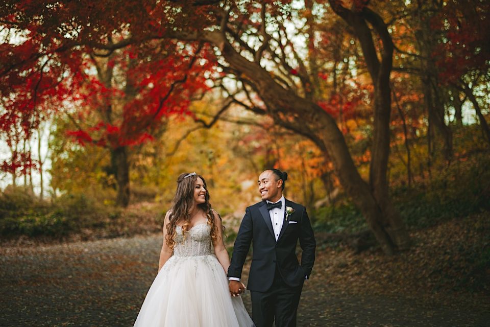 A bride and groom smiling at each other on a forest path surrounded by vibrant autumn foliage at the Ash + John Boathouse.