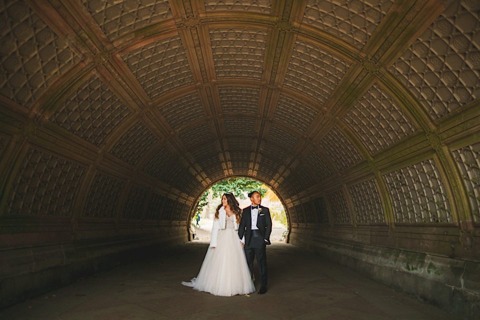 A bride and groom holding hands in an arched stone tunnel at the Ash + John Boathouse NYC Wedding, illuminated by soft light, both smiling and dressed in wedding attire.