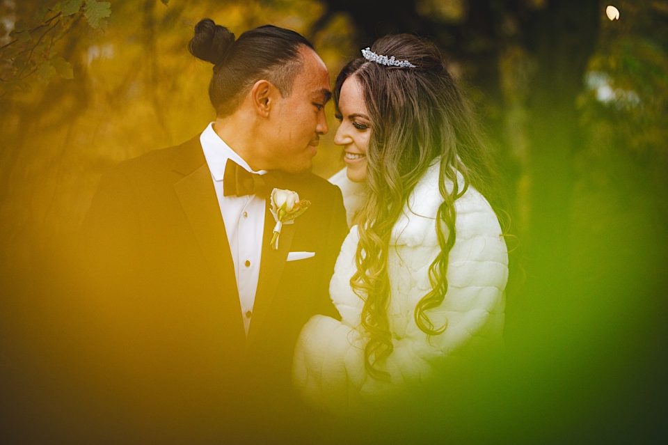 Bride and groom sharing an intimate moment at the Ash + John Boathouse, framed by soft-focus greenery, with the groom in a classic tuxedo and the bride in a white gown and tiara.