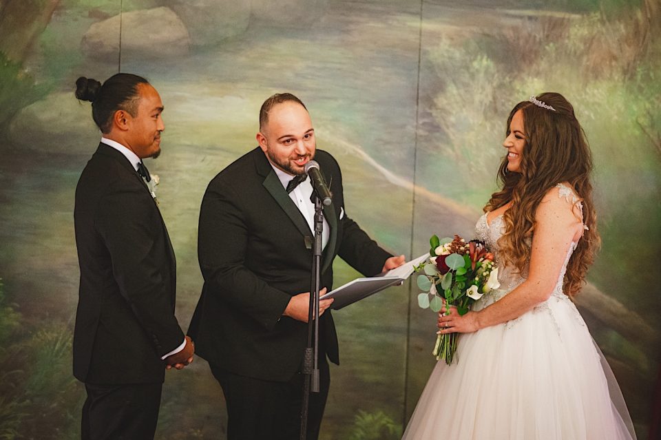 A wedding officiant speaks at a microphone during an Ash + John Boathouse NYC Wedding, standing between a smiling groom and a bride holding a bouquet.