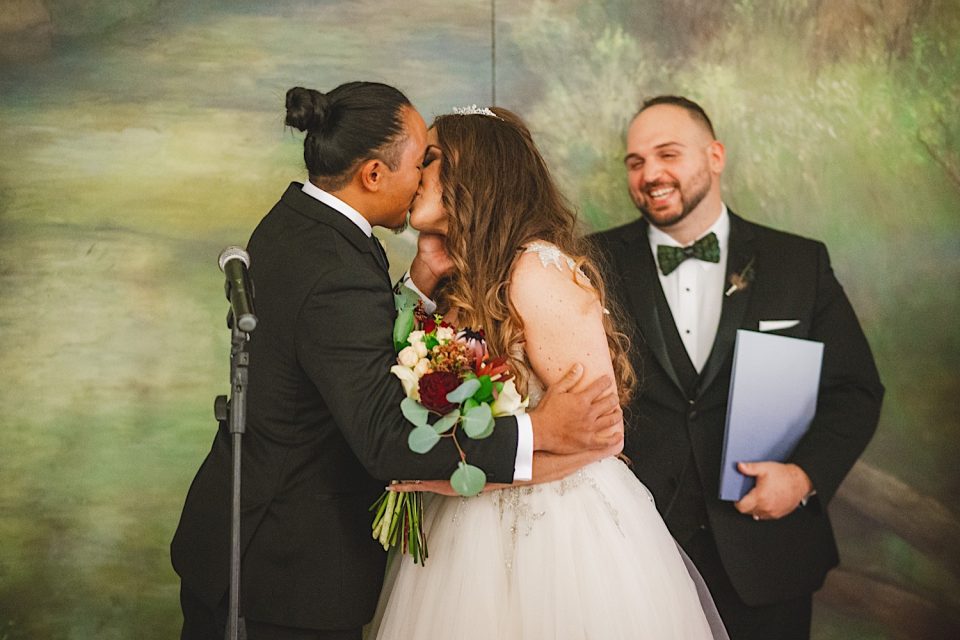A bride and groom kissing while an officiant smiles approvingly, standing in front of a painted backdrop at a Boathouse Wedding in NYC.