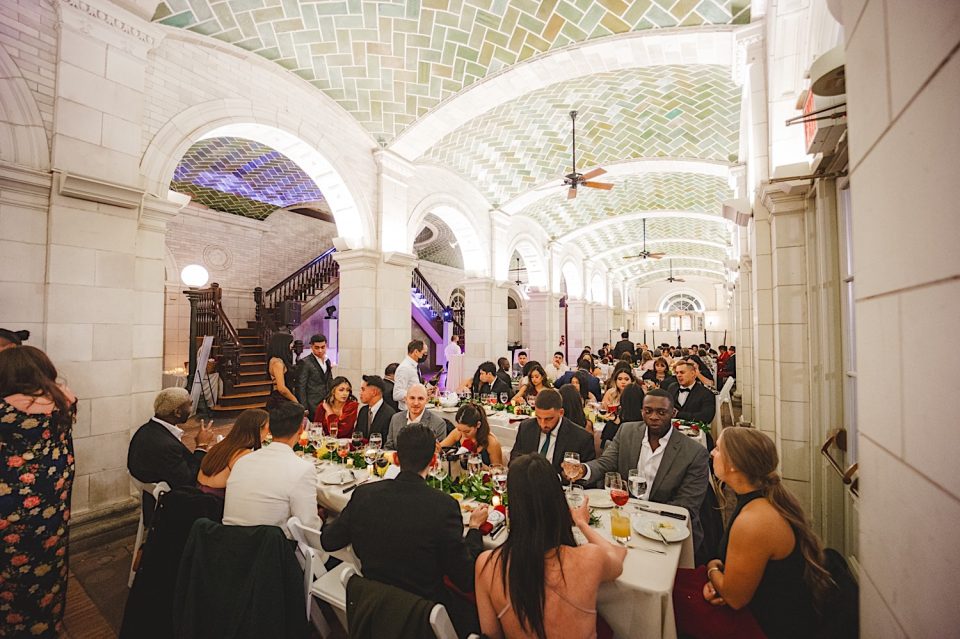 Guests seated at round tables enjoying a meal in an elegantly arched, tiled hall during a formal NYC wedding.